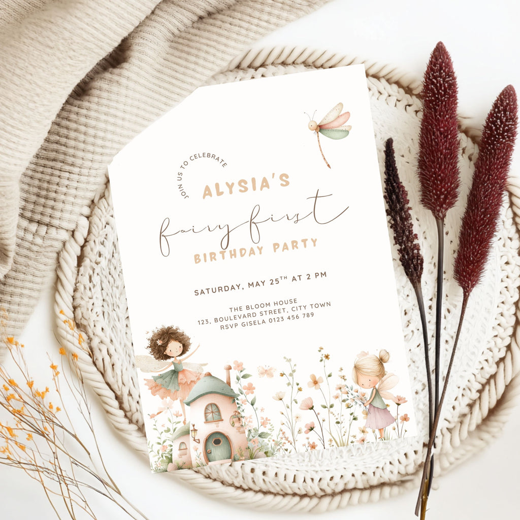 A fairy-themed first birthday party invitation card styled in a flat lay on a woven placemat with a beige knit blanket, dried bunny tail grass, and small yellow flowers. The invitation features pastel illustrations of fairies, a dragonfly, and a small cottage.