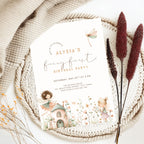 A fairy-themed first birthday party invitation card styled in a flat lay on a woven placemat with a beige knit blanket, dried bunny tail grass, and small yellow flowers. The invitation features pastel illustrations of fairies, a dragonfly, and a small cottage.