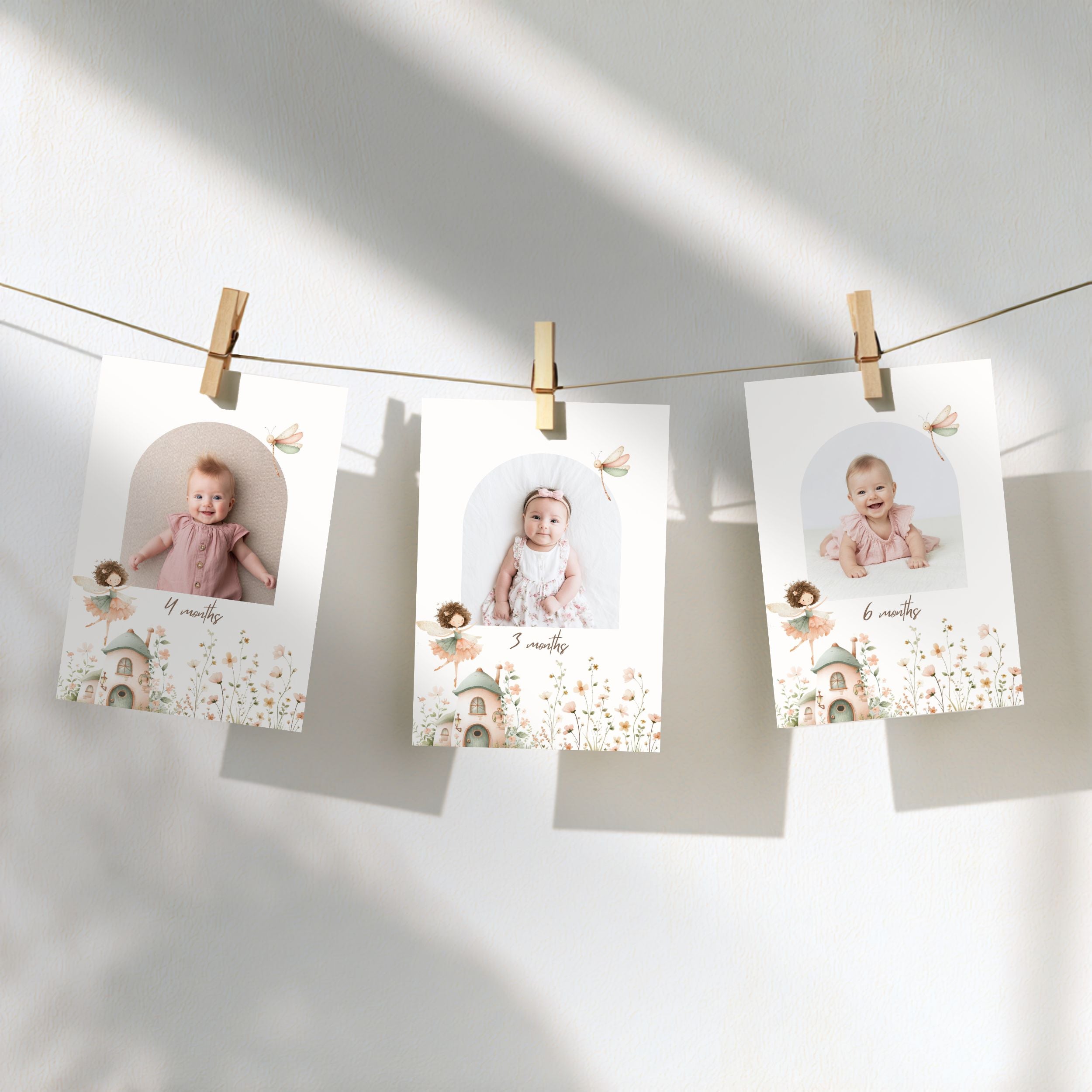 Three personalized baby milestone photos clipped to a string against a sunlit white wall, with soft shadows and delicate floral illustrations on the cards.