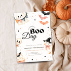 A Halloween-themed first birthday invitation card styled in a flat lay with pumpkins, fabric, and autumn leaves. The white card features cute illustrations of ghosts, bats, a witch, and a cupcake, with the text 'It's Jennifer's Boo Day' and event details.