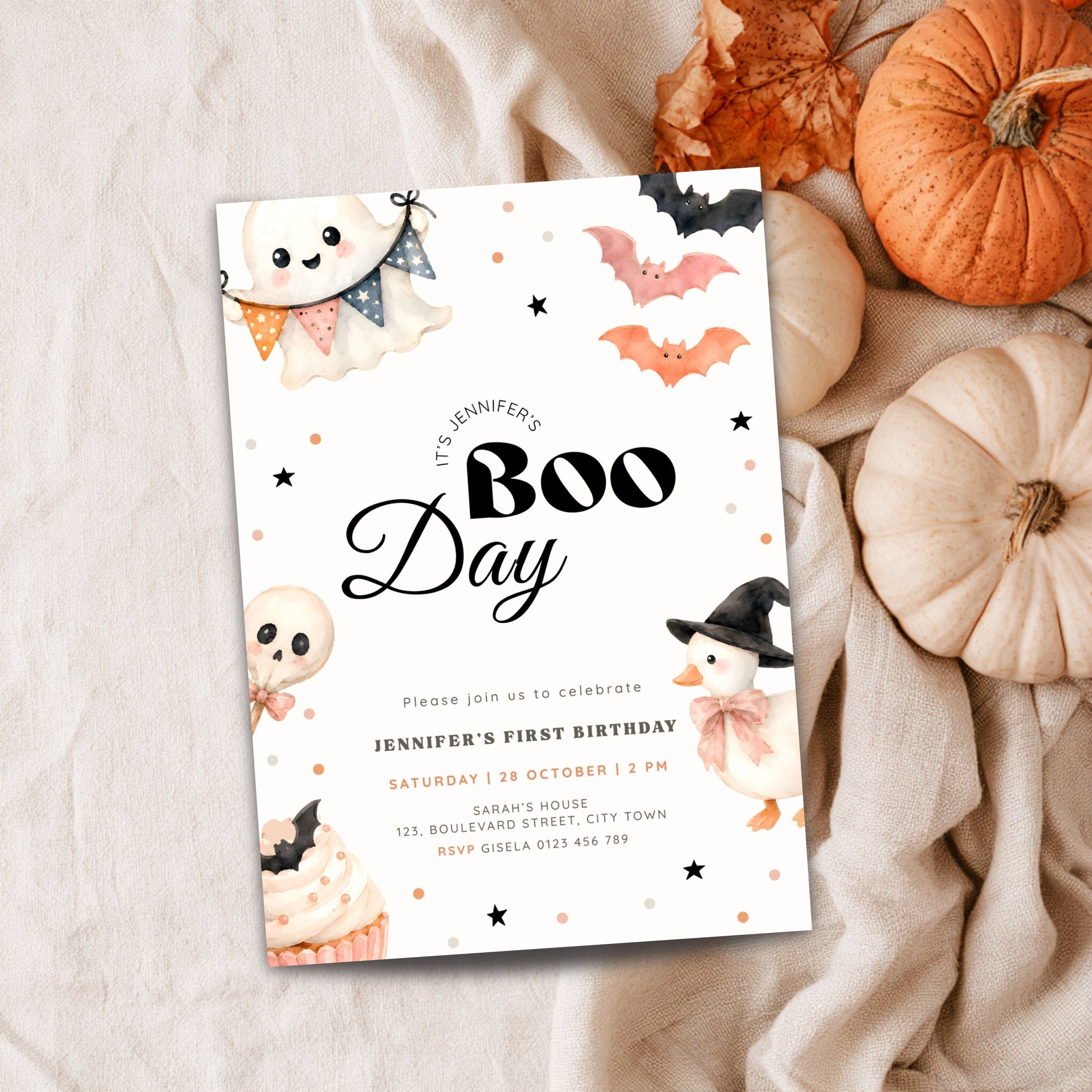 A Halloween-themed first birthday invitation card styled in a flat lay with pumpkins, fabric, and autumn leaves. The white card features cute illustrations of ghosts, bats, a witch, and a cupcake, with the text 'It's Jennifer's Boo Day' and event details.