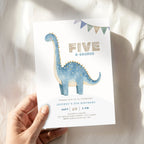 A hand holds a children's 5th birthday invitation card featuring a blue watercolor dinosaur and the text 'FIVE A-SAUROS' on a soft white fabric background with natural shadows.
