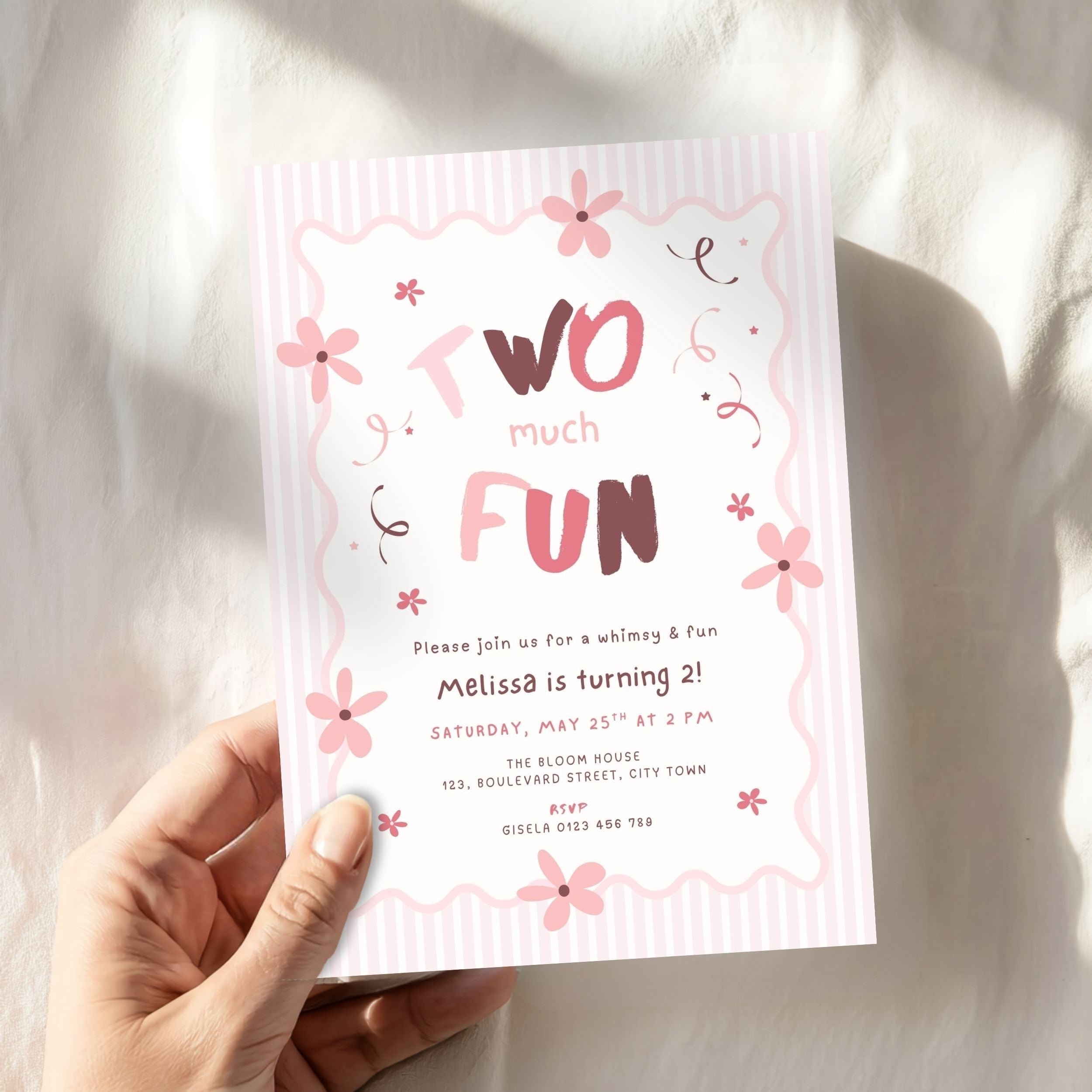 A hand holding a pink and white birthday invitation card that reads 'Two much FUN' with floral illustrations, set against a soft white fabric background with natural shadows.