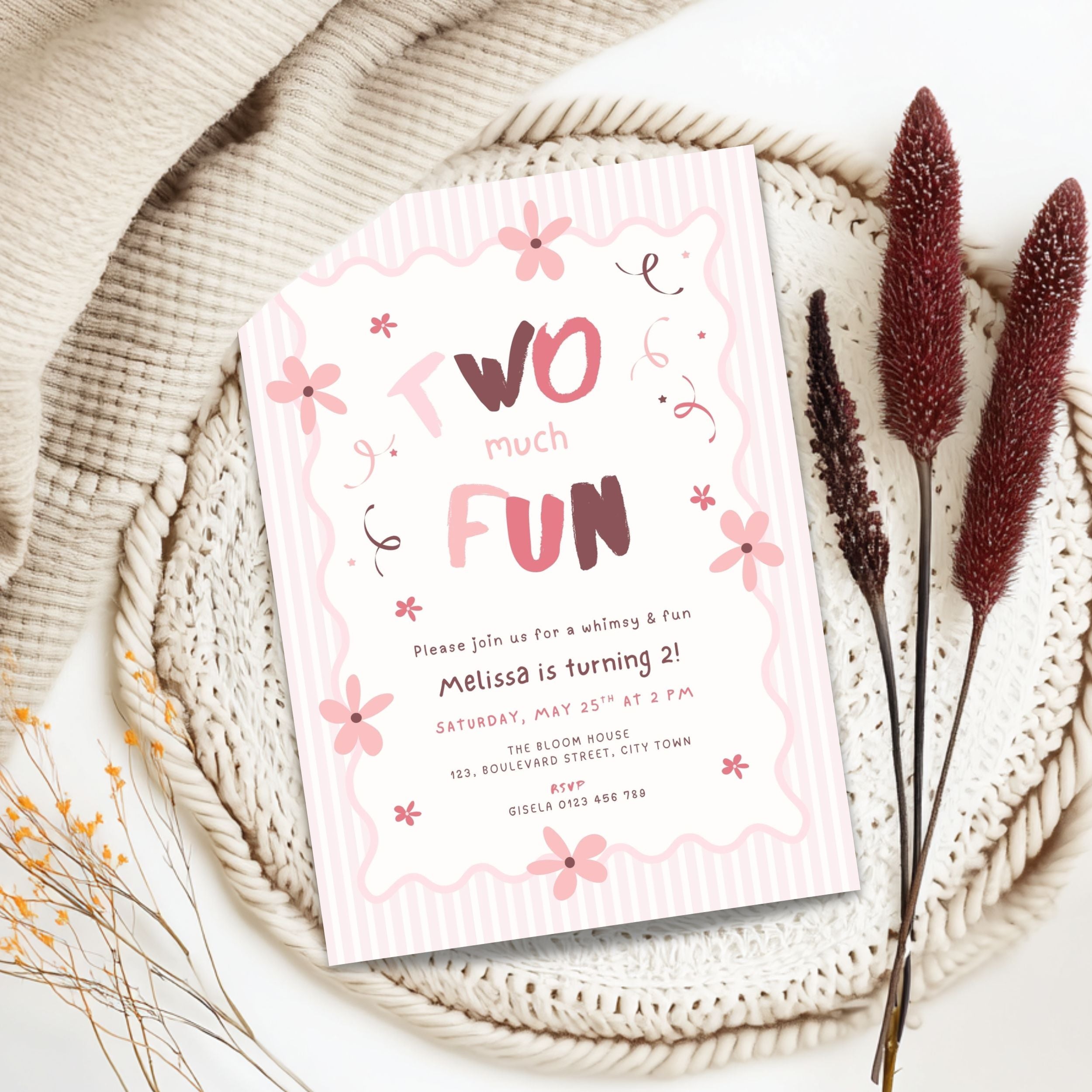 A two-year birthday invitation card styled in a flat lay on a woven placemat with dried decorative grass stems. The card is white with pink wavy borders, pink flowers, and pink and dark red lettering that reads 'Two much FUN'.