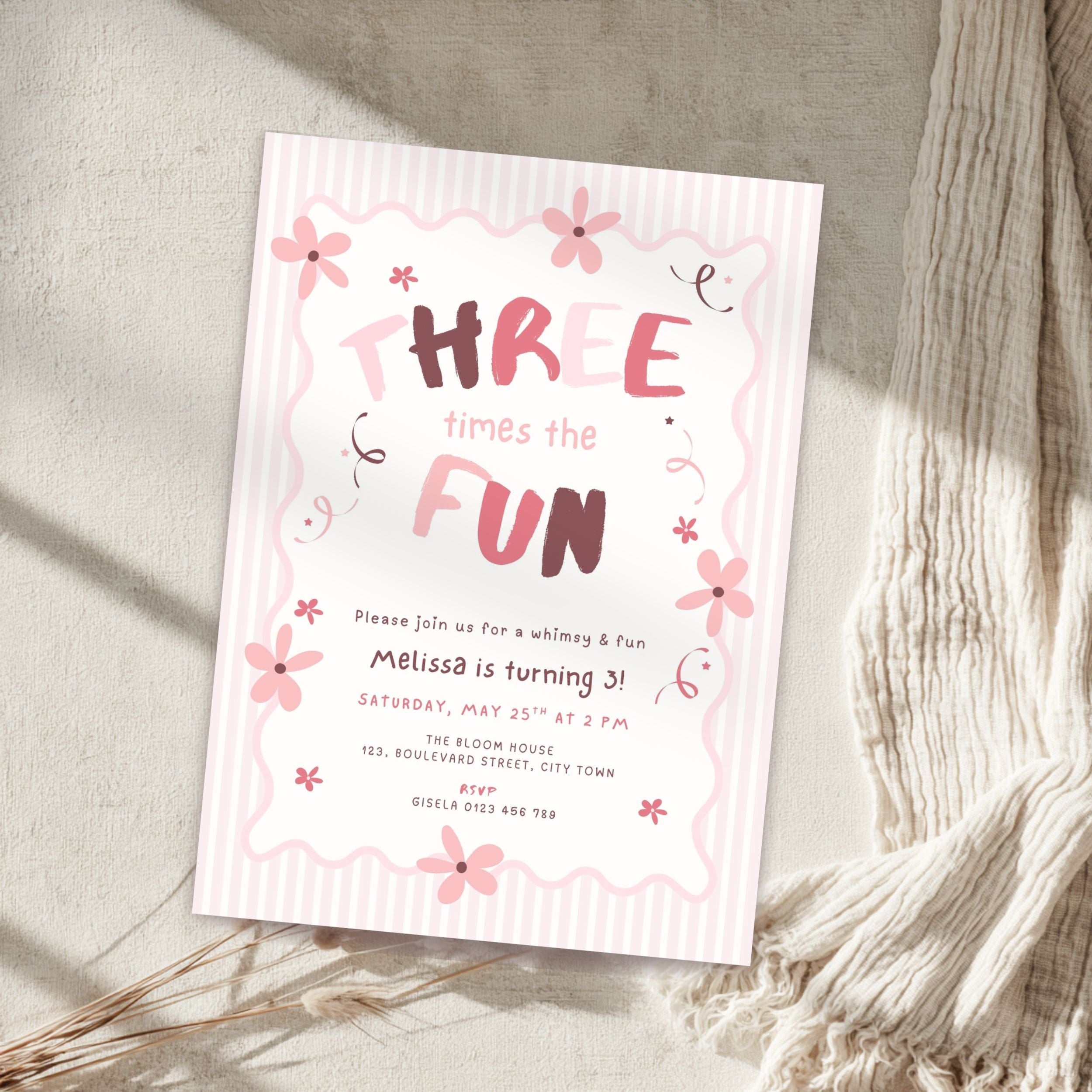 A pink and white striped three-year birthday invitation card styled on a textured beige surface with a cream knit blanket and dried grasses.