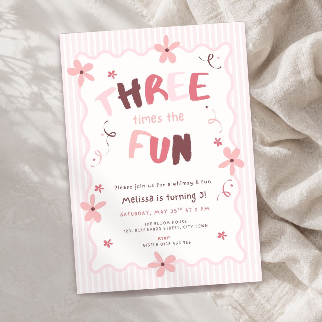 A pink and white striped 3rd birthday invitation card with floral illustrations and the text 'THREE times the FUN' in stylized lettering, photographed on a soft beige fabric background.
