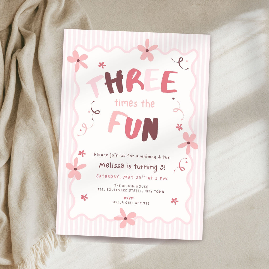 A pink and white striped 3rd birthday invitation card with floral accents, styled on a beige fabric surface next to a draped cream throw blanket.