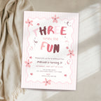 A pink and white striped 3rd birthday invitation card with floral accents, styled on a beige fabric surface next to a draped cream throw blanket.
