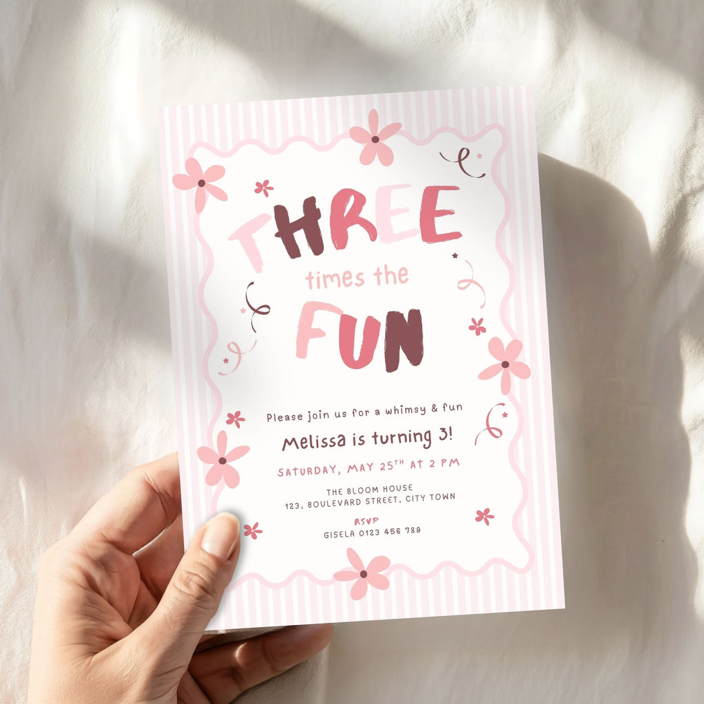 A hand holding a pink and white striped 'Three Times the Fun' birthday invitation card with floral illustrations, set against a soft white fabric background with natural shadows.