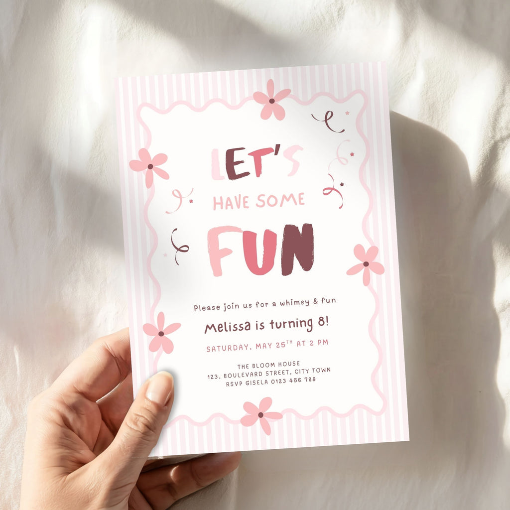 A hand holds a pink and white birthday invitation card that reads 'LET'S HAVE SOME FUN' with floral illustrations, set against a softly lit white fabric background.