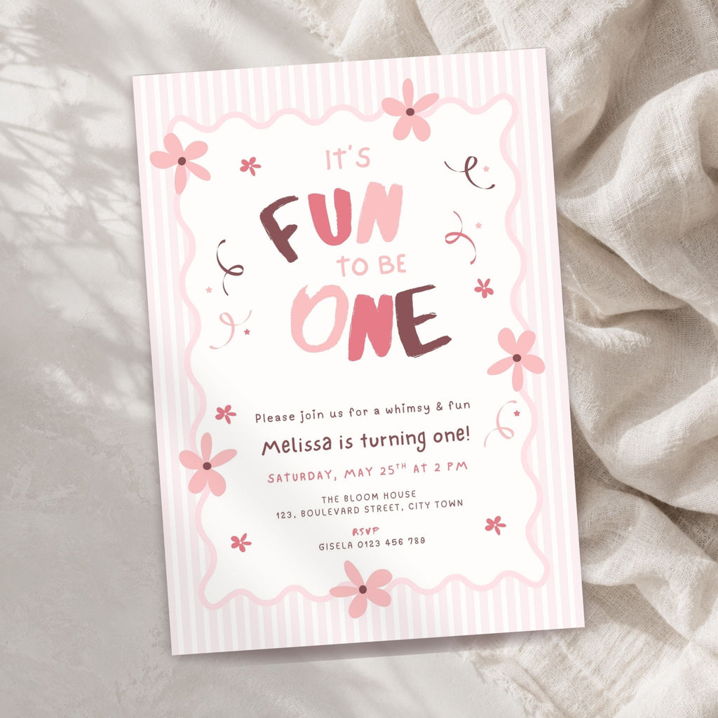 A pink and white striped baby first birthday invitation card with floral illustrations, styled on a textured beige fabric background.
