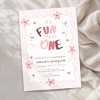A pink and white striped baby first birthday invitation card with floral illustrations, styled on a textured beige fabric background.