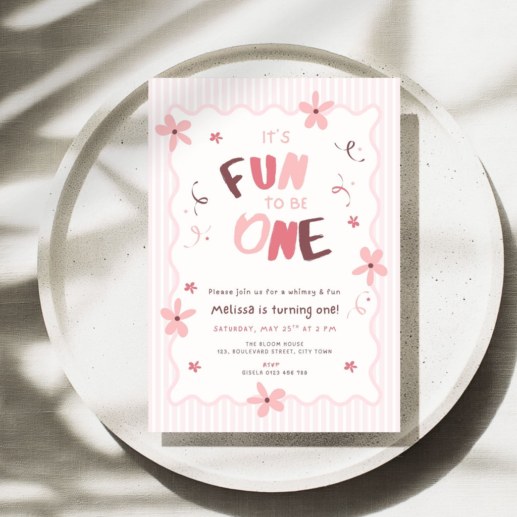 A pink and white first birthday invitation card with cute floral illustrations and the text 'It's Fun to Be One', styled on a round concrete plate with soft leaf shadows.