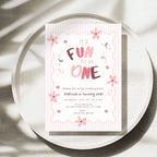 A pink and white first birthday invitation card with cute floral illustrations and the text 'It's Fun to Be One', styled on a round concrete plate with soft leaf shadows.