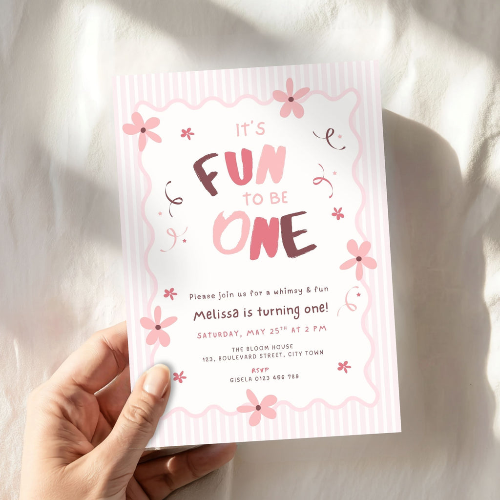 A hand holding a pink and white first birthday invitation card that reads 'It's Fun to Be One' with floral illustrations, set against a softly draped white fabric background.