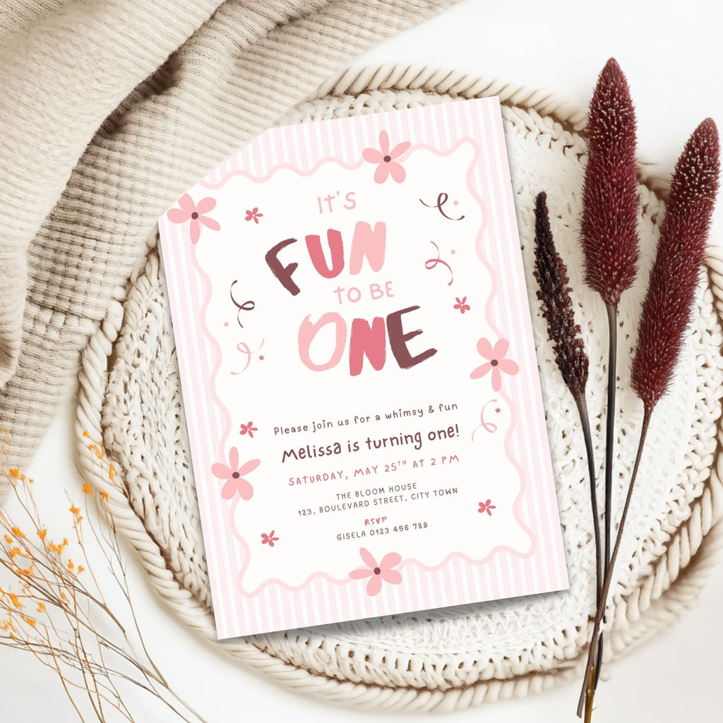 A pink and white baby first birthday invitation card styled in a flat lay on a beige knit blanket with dried grass stems.