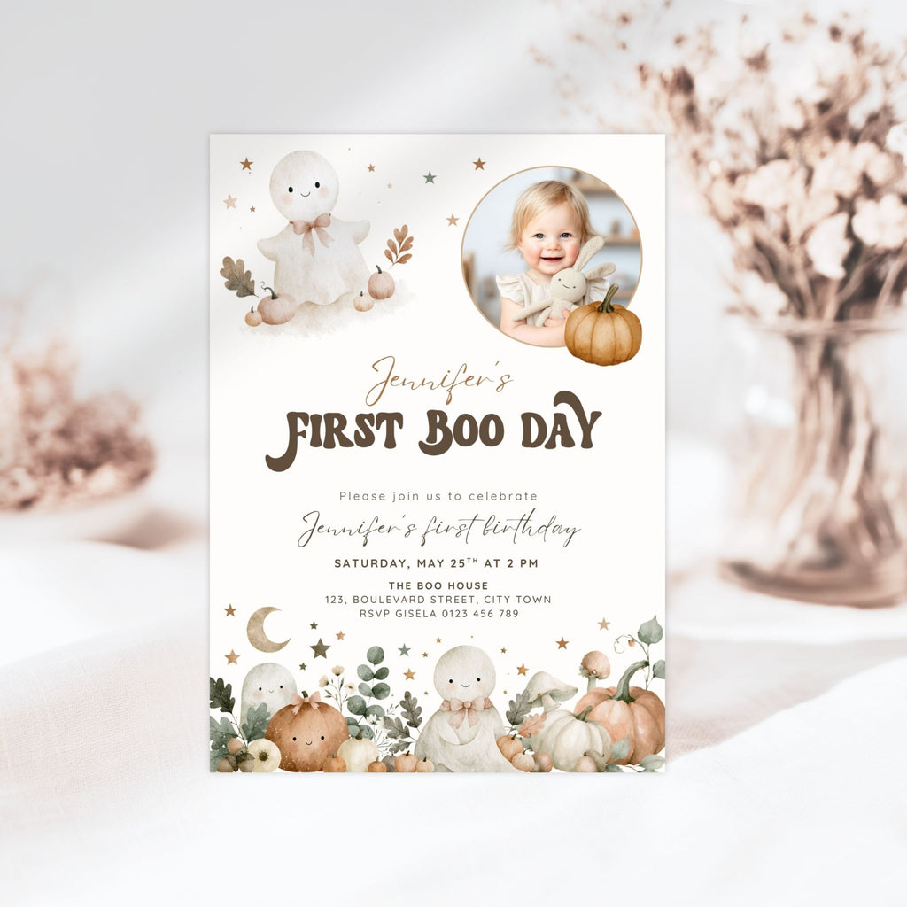 A first boo day invitation card styled in a soft, neutral setting with dried flowers in the background. The card features a white base with illustrations of cute cartoon ghosts, pumpkins, stars, and a circular photo placeholder of a baby.