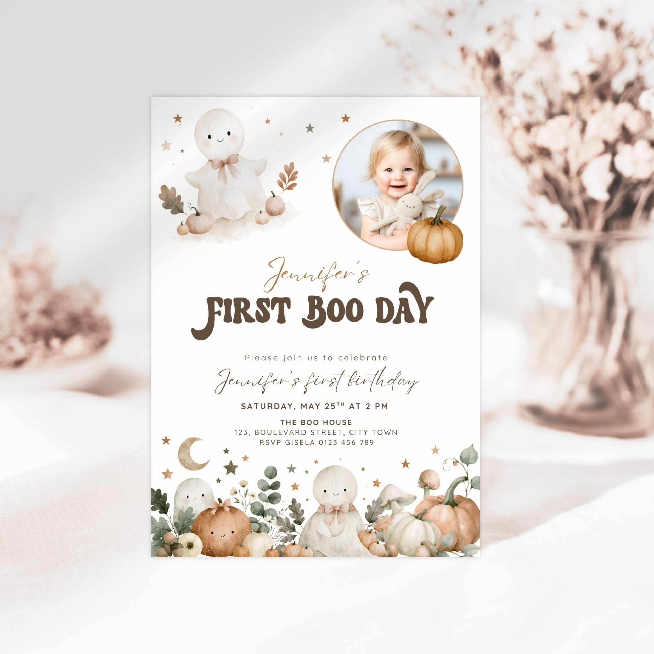 A first boo day invitation card styled in a soft, neutral setting with dried flowers in the background. The card features a white base with illustrations of cute cartoon ghosts, pumpkins, stars, and a circular photo placeholder of a baby.