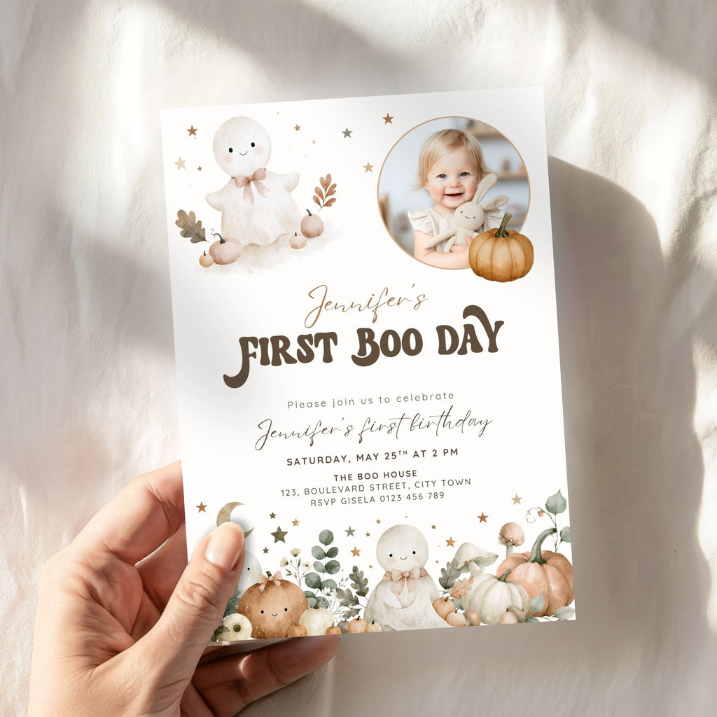 A hand holds a personalized Halloween first birthday invitation card. The card features cute watercolor illustrations of a bunny, pumpkins, and stars, with a circular photo of a young girl and the text 'Jennifer's FIRST BOO DAY'.