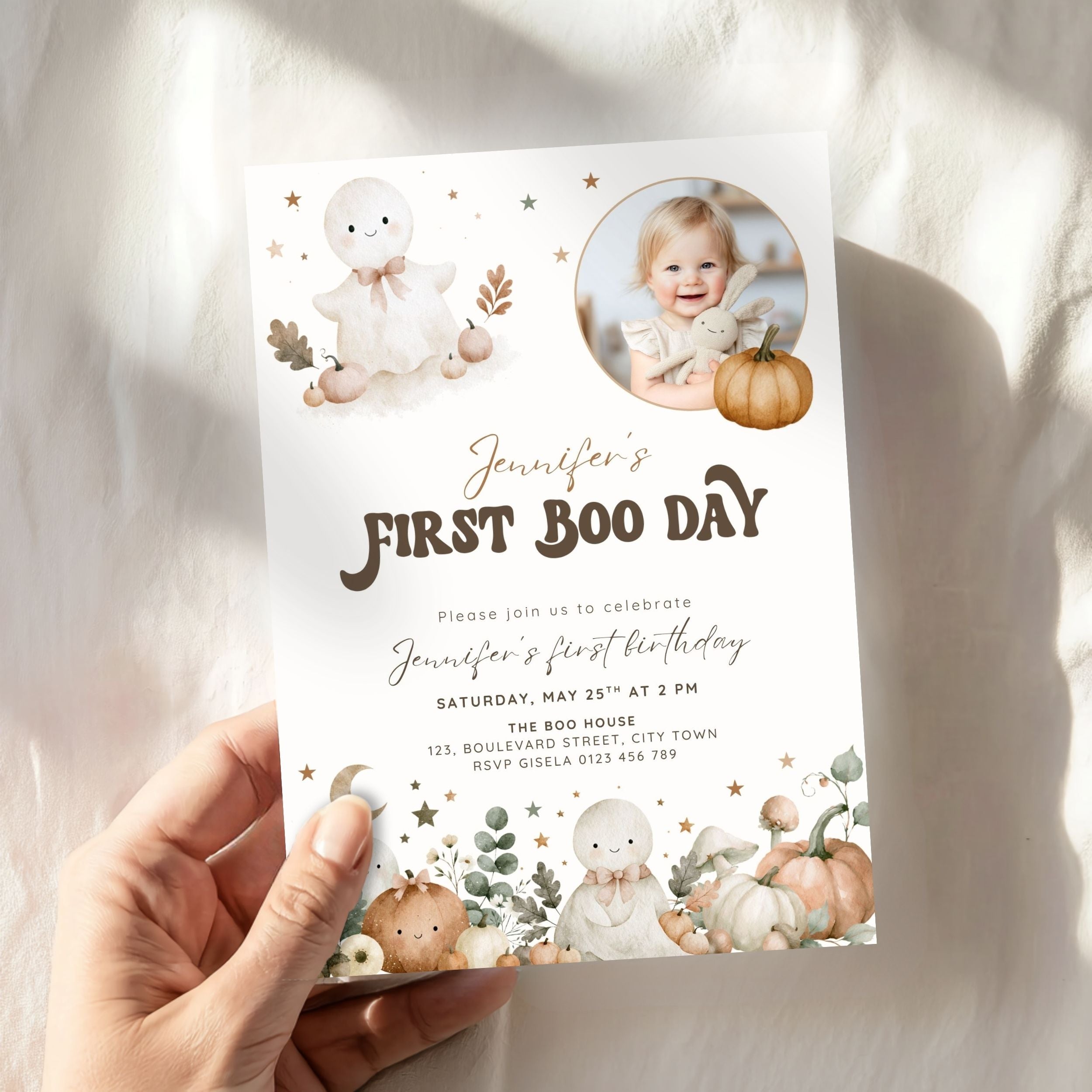 A hand holds a personalized Halloween first birthday invitation card. The card features cute watercolor illustrations of a bunny, pumpkins, and stars, with a circular photo of a young girl and the text 'Jennifer's FIRST BOO DAY'.