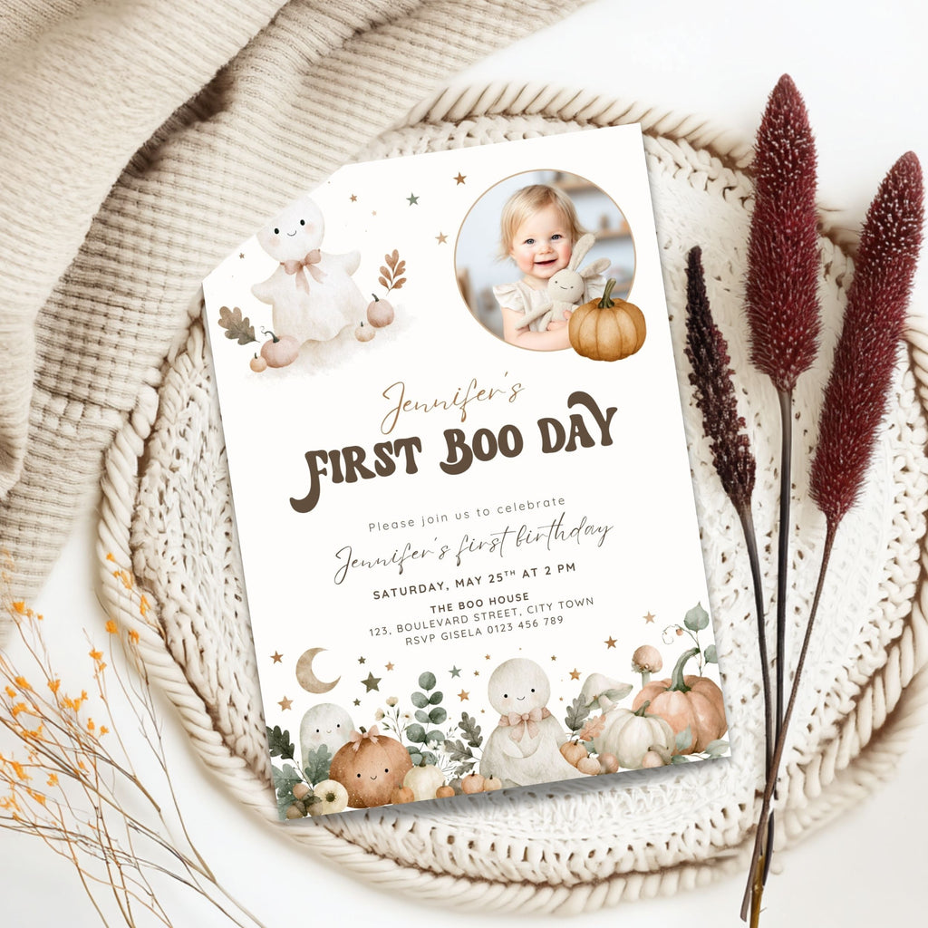 A styled flat-lay of a first birthday invitation card featuring cute watercolor baby ghosts, pumpkins, and stars, placed on a woven placemat with dried decorative grass and a beige knit blanket.