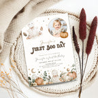 A styled flat-lay of a first birthday invitation card featuring cute watercolor baby ghosts, pumpkins, and stars, placed on a woven placemat with dried decorative grass and a beige knit blanket.