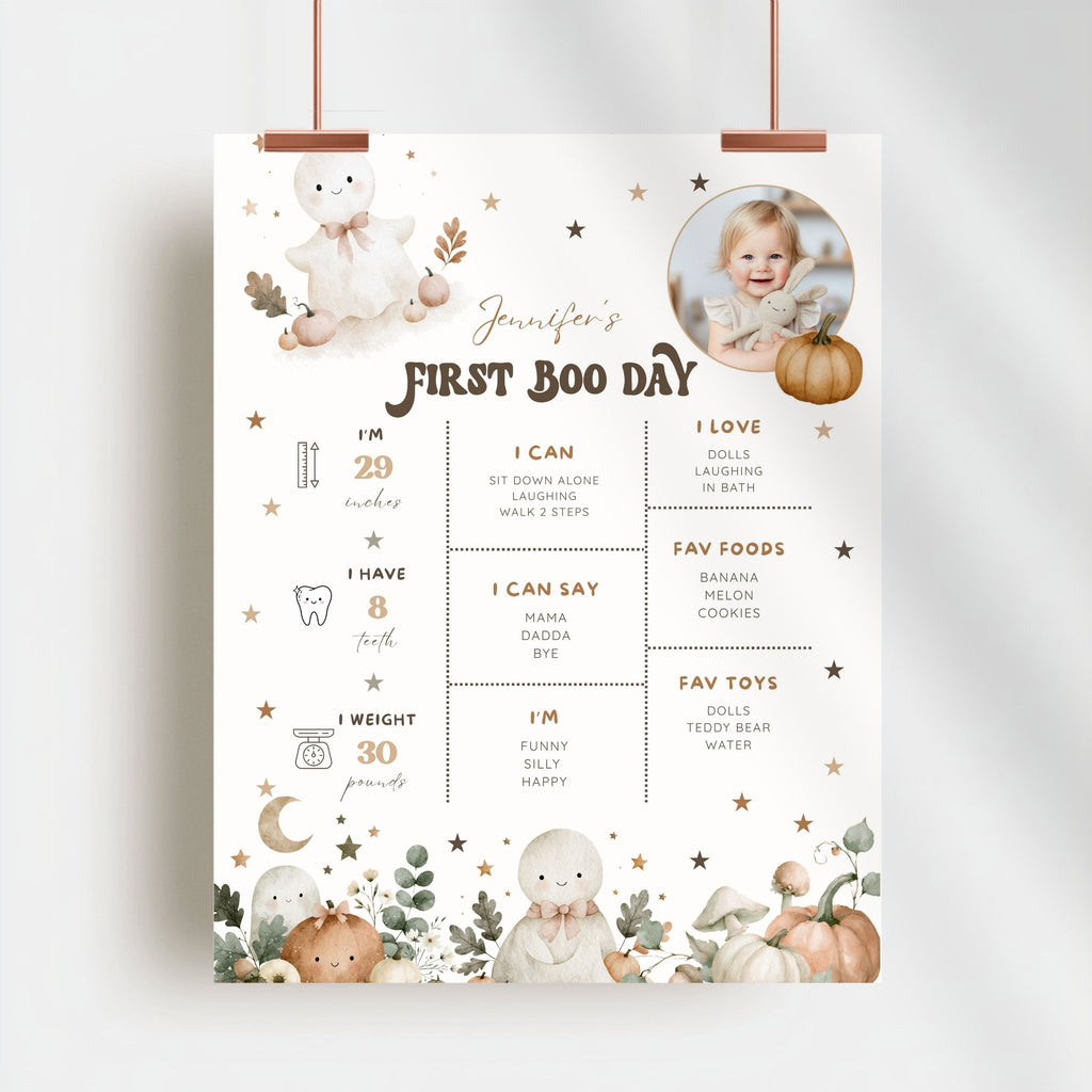A personalized first boo day milestone poster for a baby, featuring cute illustrations of bears, pumpkins, and fall leaves on a cream background. The poster includes customizable text sections for the baby's name, age, skills, foods, and toys, and is shown hanging from two copper-colored clips against a light gray wall.