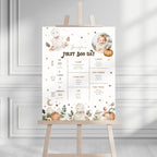 A baby first birthday party sign displayed on a wooden easel. The sign features a warm, neutral color palette with illustrations of a teddy bear, pumpkins, stars, and a baby photo, alongside sections for baby milestones, favorite foods, and toys.