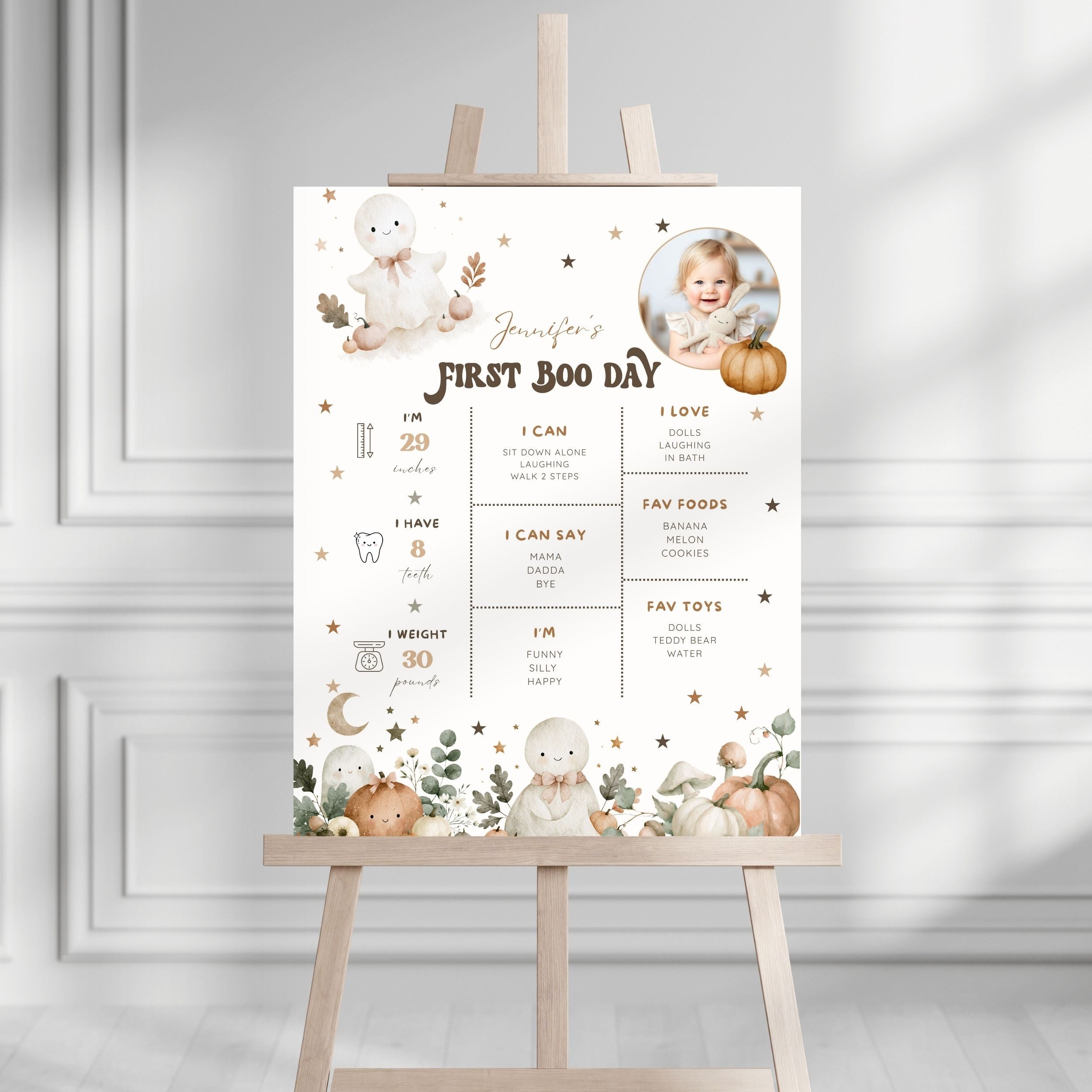A baby first birthday party sign displayed on a wooden easel. The sign features a warm, neutral color palette with illustrations of a teddy bear, pumpkins, stars, and a baby photo, alongside sections for baby milestones, favorite foods, and toys.