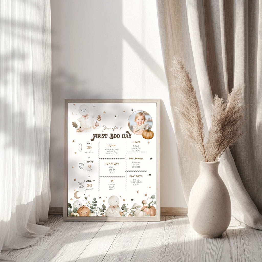 A framed baby first birthday milestone chart with cute watercolor illustrations of a baby, pumpkins, and woodland motifs, styled on a light wood floor next to a vase with pampas grass in a sunlit room.