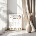 A framed baby first birthday milestone chart with cute watercolor illustrations of a baby, pumpkins, and woodland motifs, styled on a light wood floor next to a vase with pampas grass in a sunlit room.