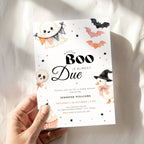 A hand holds a Halloween baby shower invitation card with cute illustrations of a ghost, witch chick, balloons, bats, and a cupcake against a soft white fabric background.