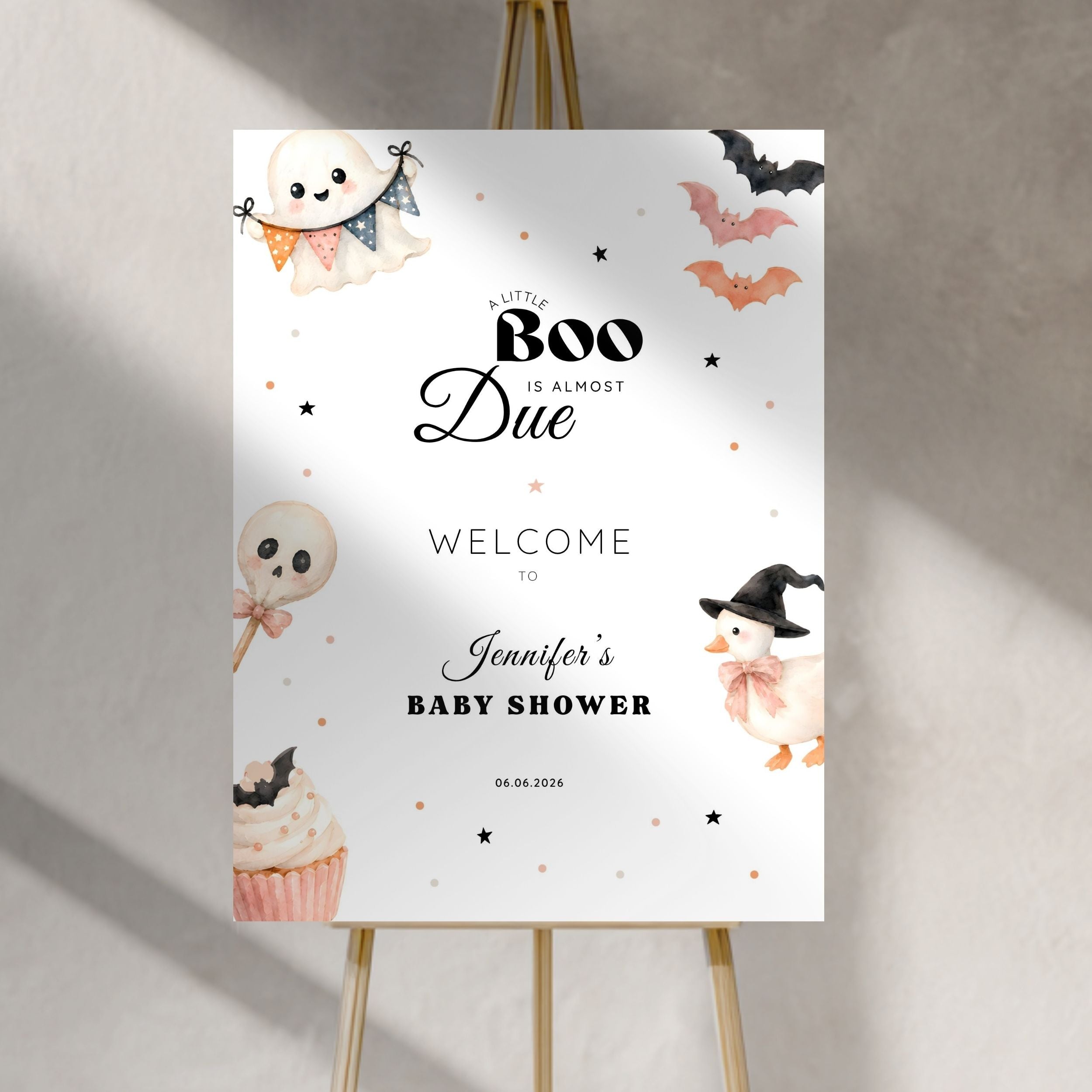 A vertical white baby shower sign with Halloween illustrations, displayed on a gold easel against a textured gray wall with soft shadows.