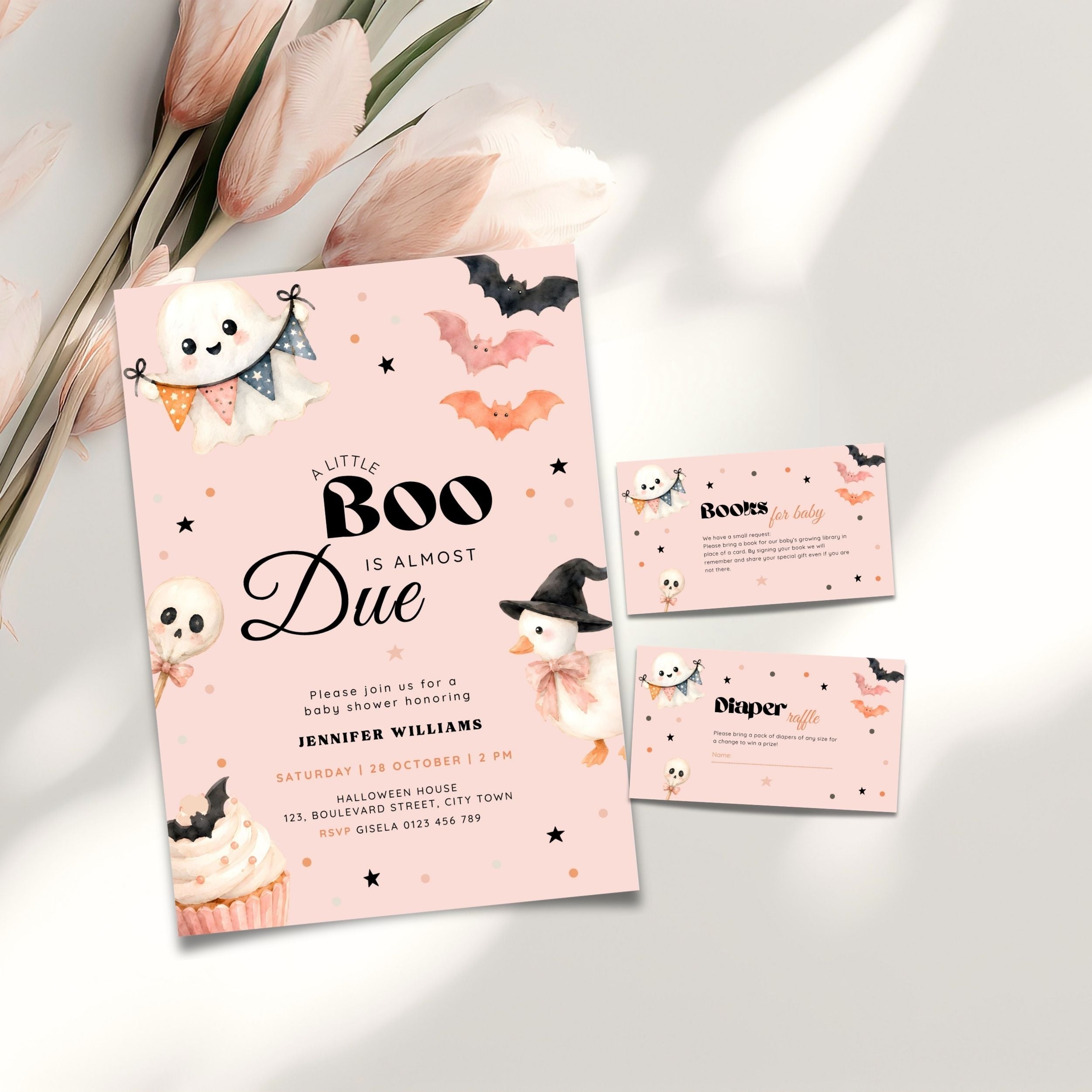A Halloween baby shower invitation set with a light blue card and matching envelopes, decorated with cartoon ghosts, bats, cupcakes, and a witch, styled on a white surface with pink tulips.