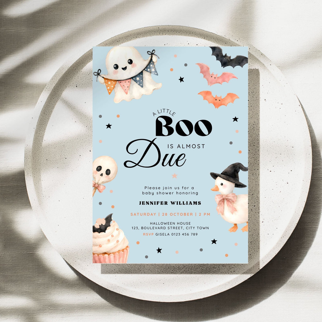 A Halloween-themed baby shower invitation card displayed on a round tray. The card has a light blue background with cute illustrations of a ghost, bats, an orange pumpkin, a witch duck, a cupcake, and black decorative stars.