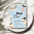 A Halloween-themed baby shower invitation card displayed on a round tray. The card has a light blue background with cute illustrations of a ghost, bats, an orange pumpkin, a witch duck, a cupcake, and black decorative stars.
