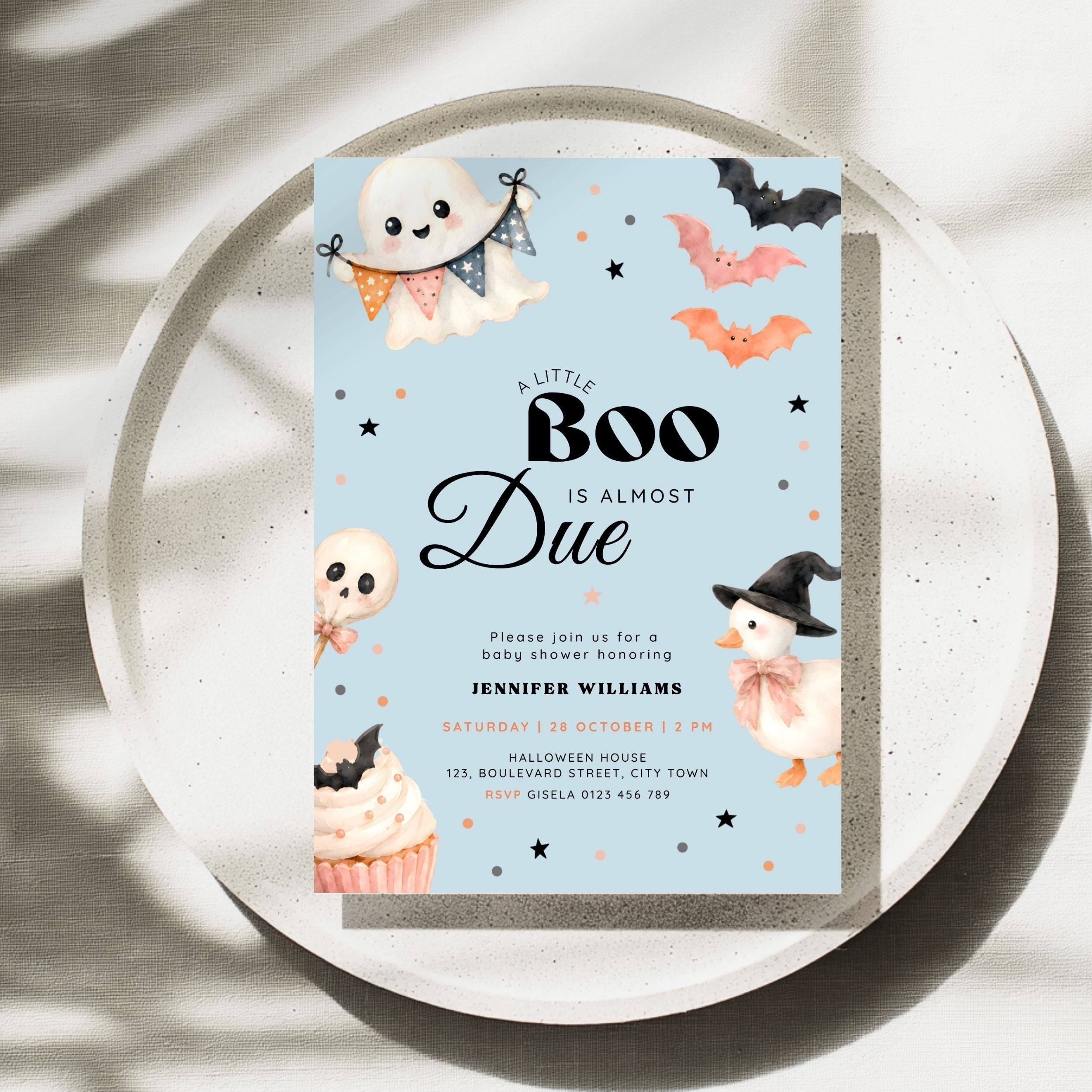A Halloween-themed baby shower invitation card displayed on a round tray. The card has a light blue background with cute illustrations of a ghost, bats, an orange pumpkin, a witch duck, a cupcake, and black decorative stars.