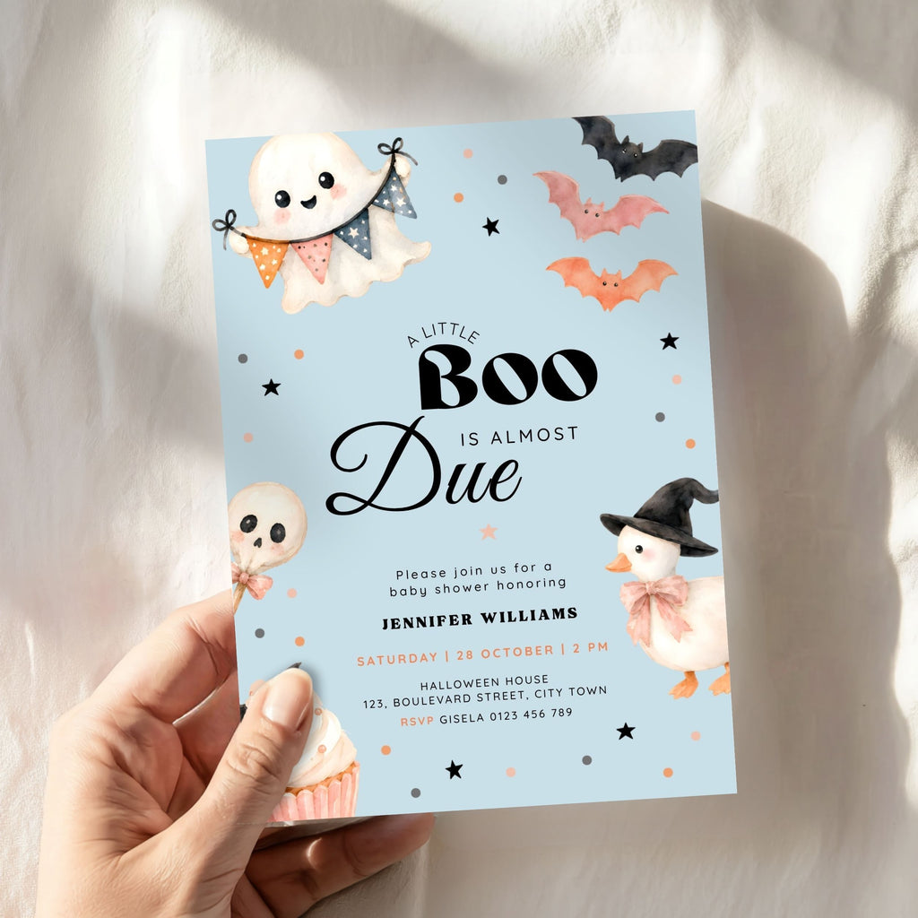 A hand holds a Halloween baby shower invitation card with a light blue background, featuring cute illustrations of a ghost, witch duck, bats, and cupcakes. The card is photographed against a soft white fabric backdrop with warm sunlight and shadows.