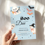 A hand holds a Halloween baby shower invitation card with a light blue background, featuring cute illustrations of a ghost, witch duck, bats, and cupcakes. The card is photographed against a soft white fabric backdrop with warm sunlight and shadows.
