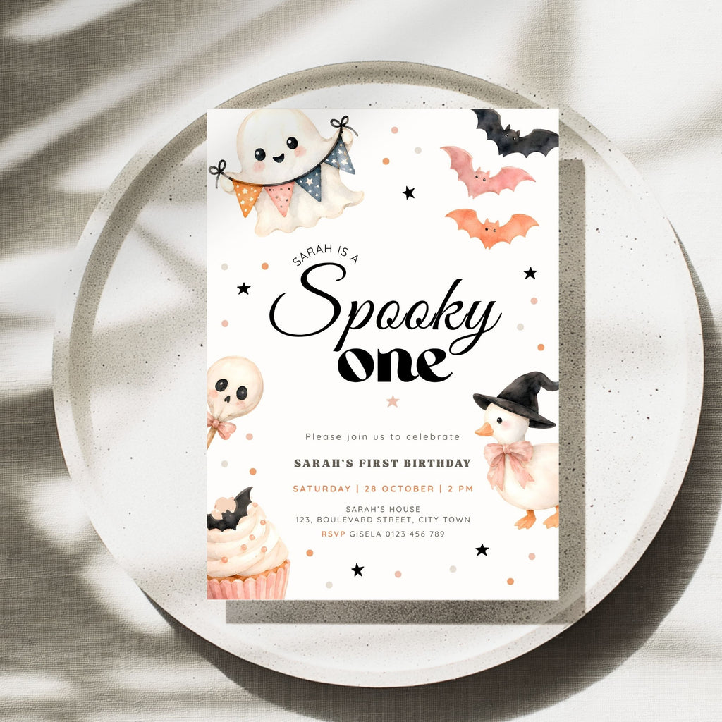 A Halloween-themed first birthday invitation card displayed on a speckled white plate with soft shadows. The card features cute illustrations of a ghost, pumpkin, witch cat, cupcake, bats, and stars, with the text 'Sarah is a Spooky one' in large black script.