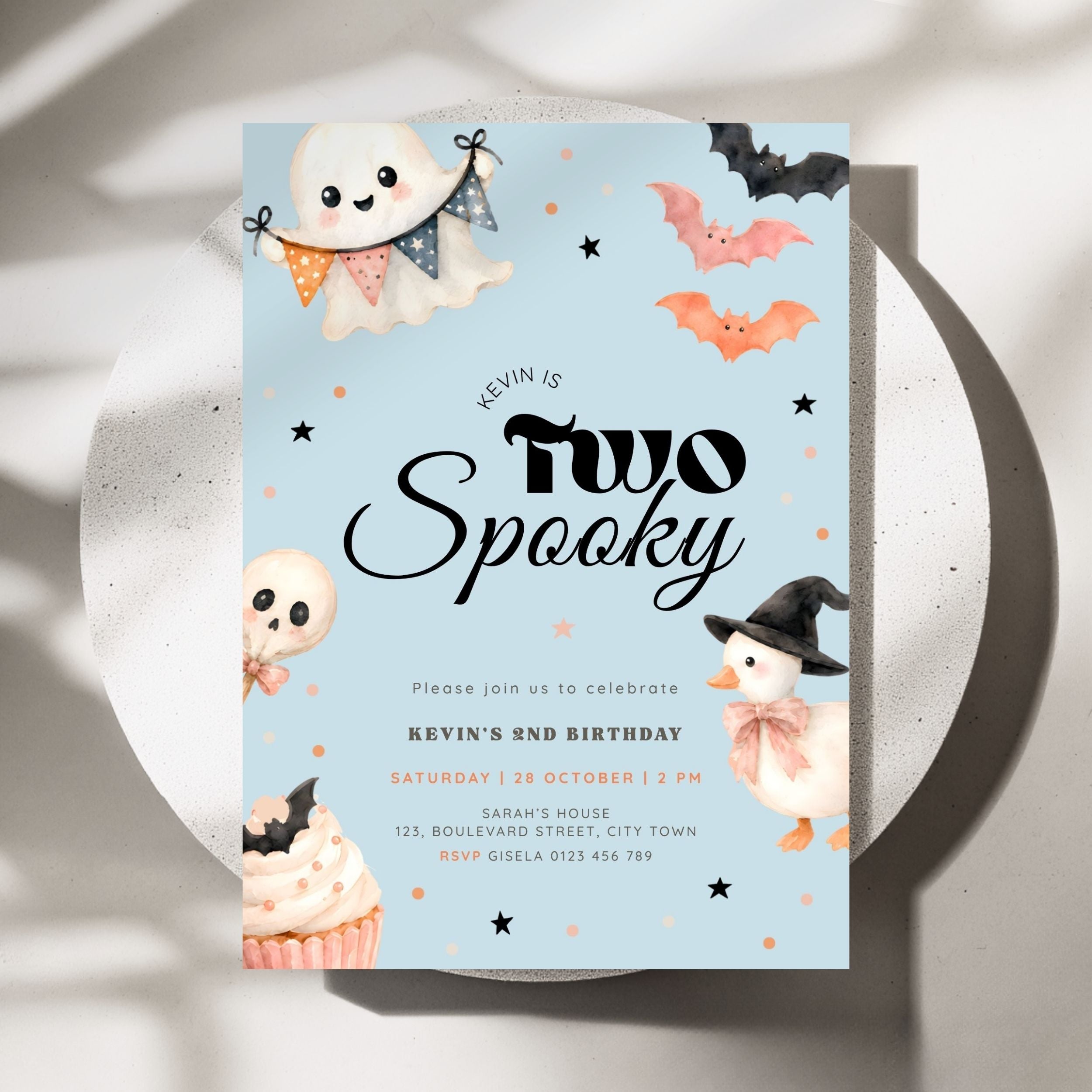A square Halloween-themed birthday invitation card with a light blue background, featuring cartoon ghosts, bats, a skull, a turkey, and a cupcake. The card reads 'KEVIN IS Two Spooky' and includes birthday event details.
