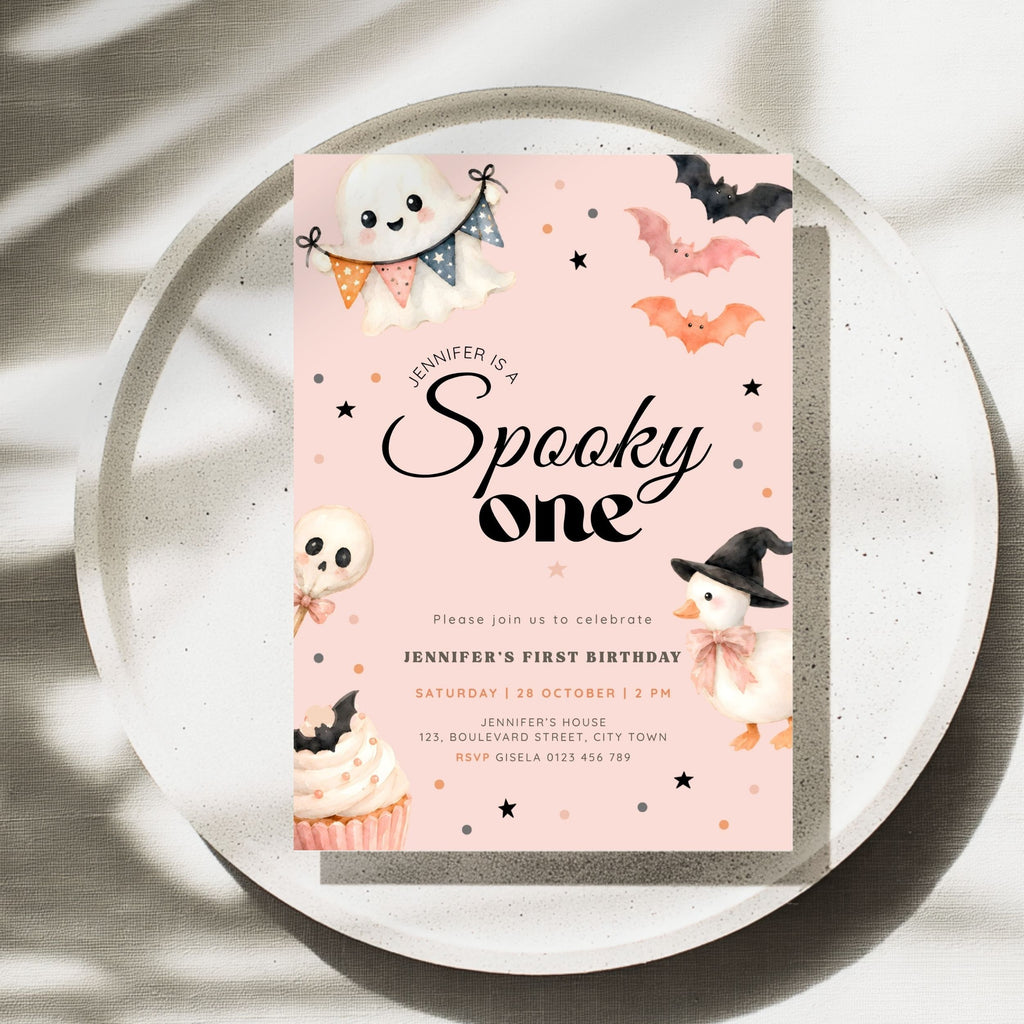 A pastel pink Halloween-themed birthday invitation card with cute illustrations of a ghost, bats, a witch, a cupcake, and stars, styled on a round white tray with soft leaf shadows.