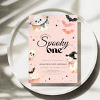 A pastel pink Halloween-themed birthday invitation card with cute illustrations of a ghost, bats, a witch, a cupcake, and stars, styled on a round white tray with soft leaf shadows.