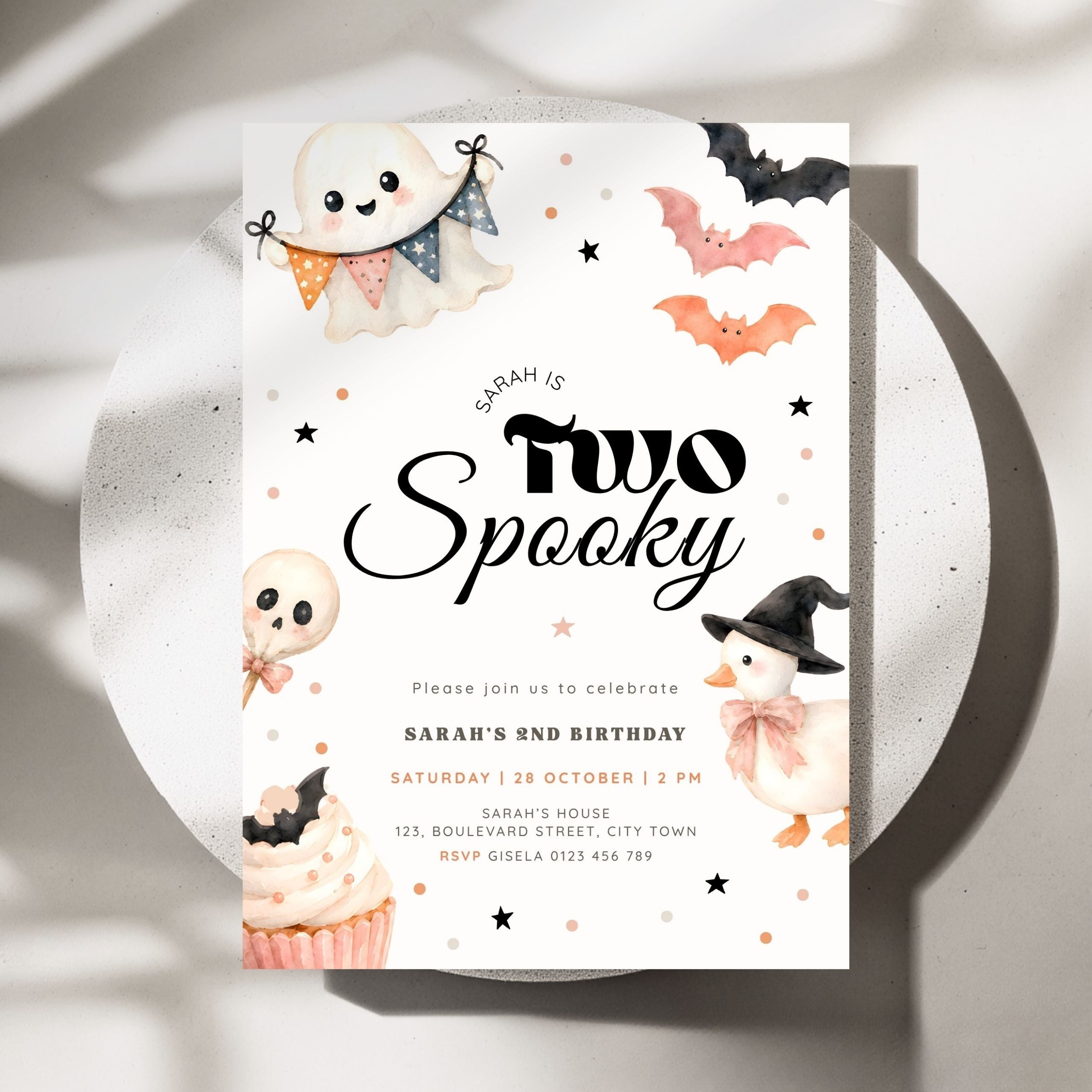 A Halloween-themed birthday invitation card for Sarah's 2nd birthday, styled on a white ceramic plate with soft shadows. The card features cute watercolor illustrations of a ghost, a pumpkin, a skull, a cupcake, and bats on a white background with black and orange text.