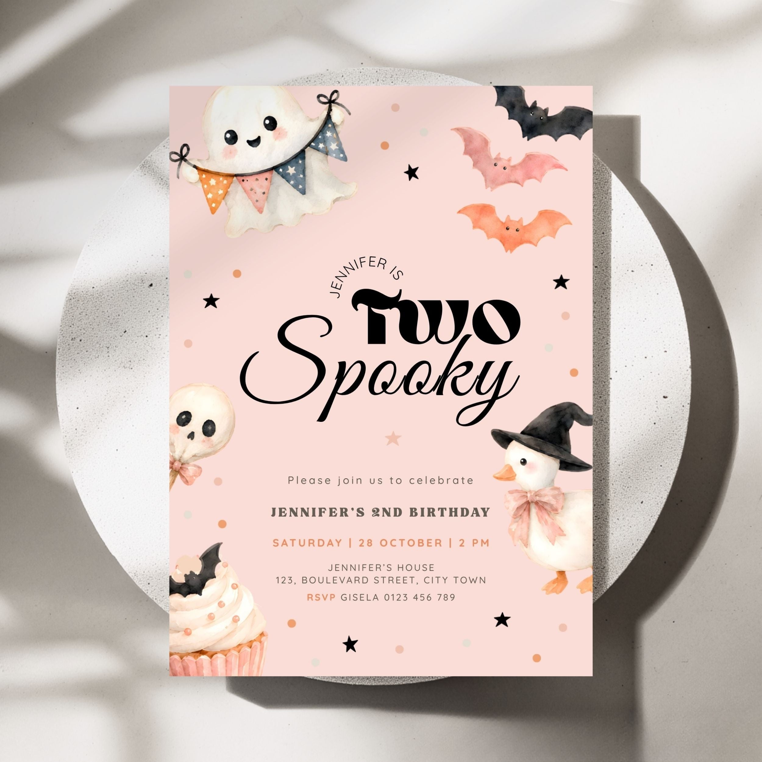 A pastel pink Halloween-themed birthday invitation card with cute illustrations of a ghost, bats, a skull, a snowman, and a cupcake, styled on a white plate with soft shadows.