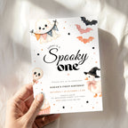 A hand holds a white Halloween-themed first birthday invitation card featuring cute cartoon illustrations of a ghost, skull, pumpkin, witch cat, bats, and stars. The card has black script text reading 'Sarah is a Spooky one' and smaller event details below, set against a softly lit white fabric background.