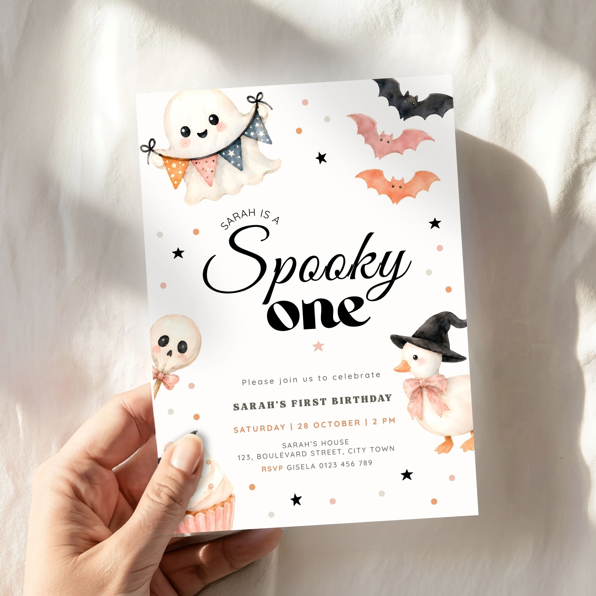 A hand holds a white Halloween-themed first birthday invitation card featuring cute cartoon illustrations of a ghost, skull, pumpkin, witch cat, bats, and stars. The card has black script text reading 'Sarah is a Spooky one' and smaller event details below, set against a softly lit white fabric background.