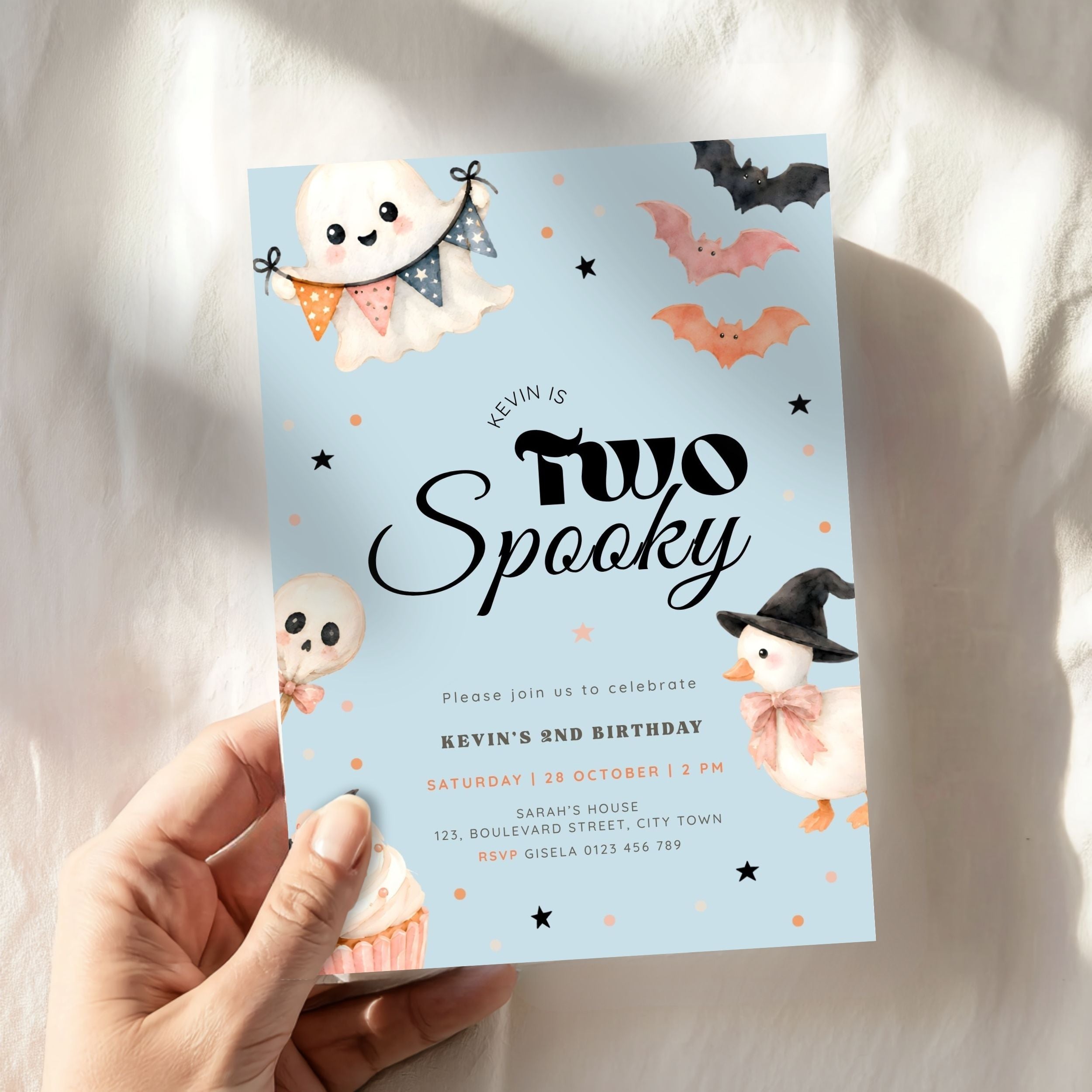 A hand holds a light blue Halloween-themed birthday invitation card that reads 'Kevin is Two Spooky' with cute ghost, bat, skull, and witch illustrations on a soft white fabric background.