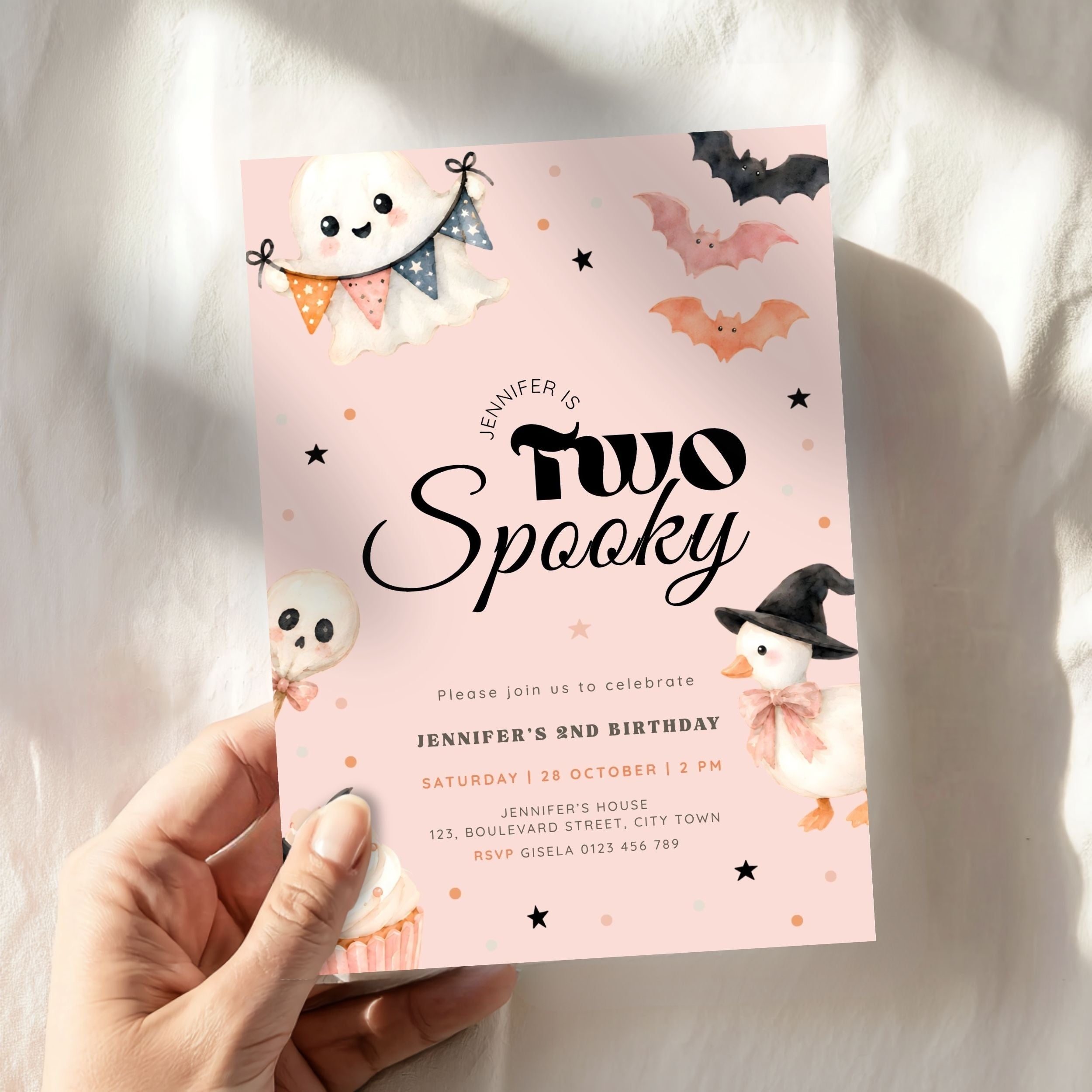 A hand holds a pink Halloween-themed birthday invitation card with cute ghost, bat, and witch illustrations. The card reads 'Jennifer is Two Spooky' and includes event details for a 2nd birthday celebration.