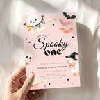 A hand holds a pink Halloween first birthday invitation card featuring cute cartoon illustrations of a ghost, witch pumpkin, bat, and skull on a soft white fabric background.